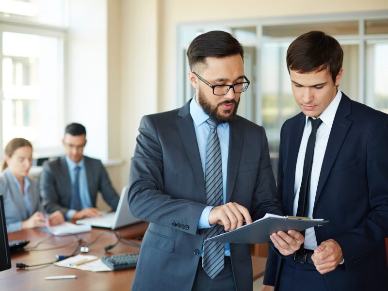 Confident businessman pointing at document while explaining his idea to his partner on background of their colleagues
