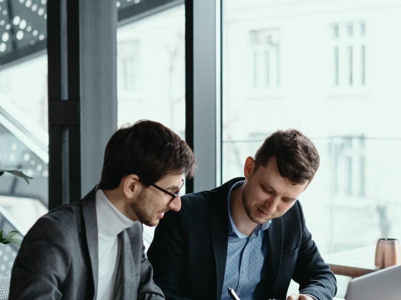 Two businessmen talking about new opportunities sitting with laptop at desk near large window, planning project, considering business offer, sharing ideas while drinking coffee together